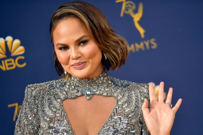 Chrissy Teigen waves at the camera, wearing a silver high-neck gown.